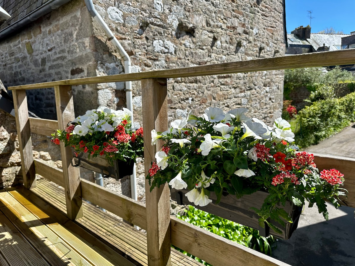 A wooden railing is adorned with flower boxes filled with white petunias and red flowers. The sunlight illuminates the blooms, while a stone wall and greenery are visible in the background, contributing to the tranquil setting.