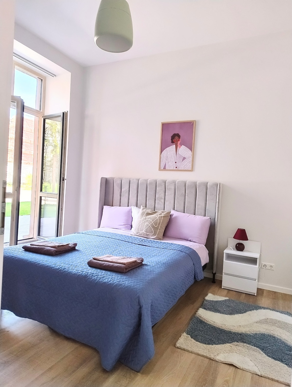 A well-appointed sleeping area features a spacious bed with a gray headboard and soft bedding in shades of blue and lavender. A bedside table holds a small lamp, while a piece of artwork adorns the wall. Large windows allow natural light to fill the room.