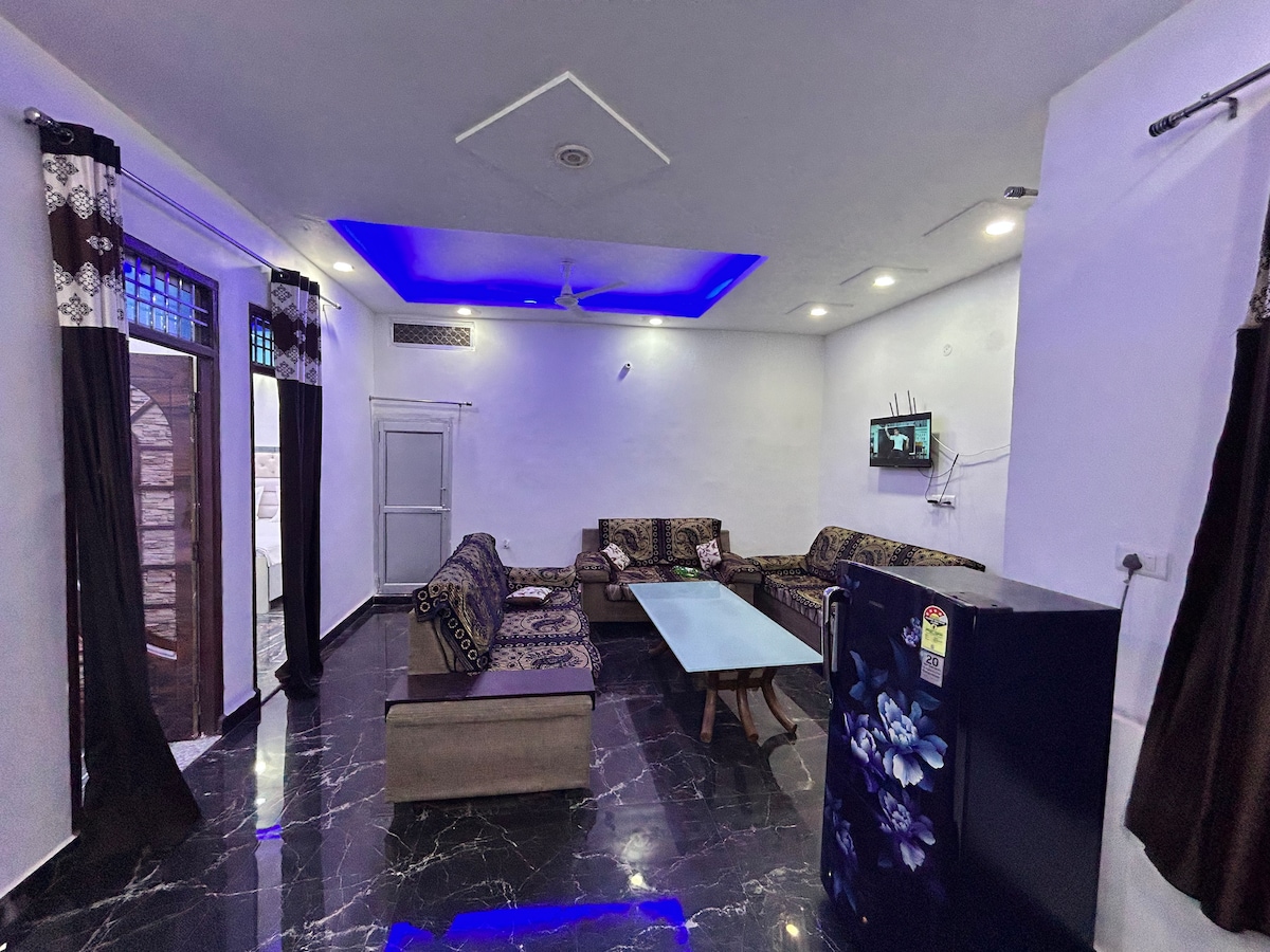 A modern living room featuring dark marble flooring and stylish furnishings. A spacious seating arrangement is present, complemented by a coffee table. A refrigerator stands in the corner, while soft lighting highlights the area. A television is mounted on the wall opposite the seating.
