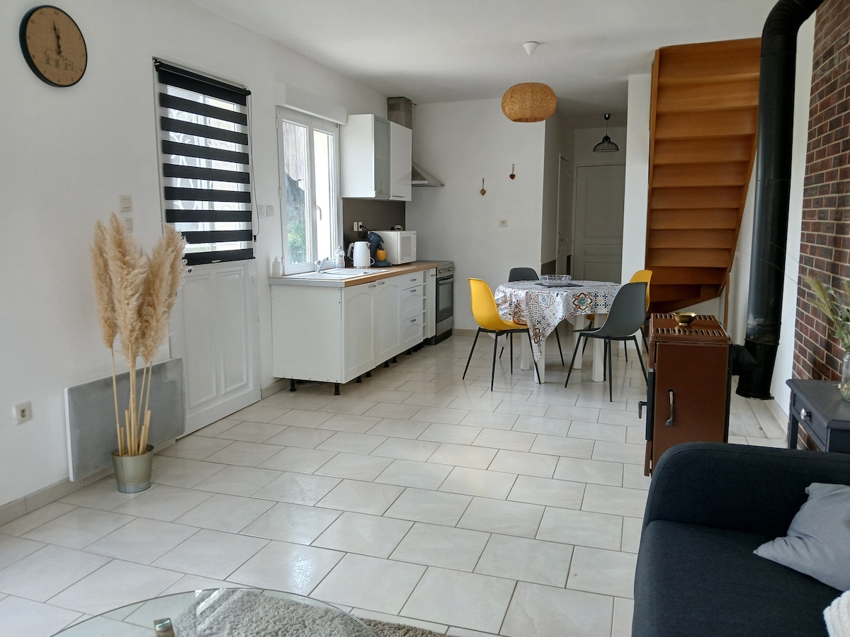 An open living space is illuminated by natural light, featuring a white kitchen with modern appliances and a dining area with a patterned tablecloth. Yellow and gray chairs surround the table, and a staircase adds visual interest. Decorative plants are placed in the corner.
