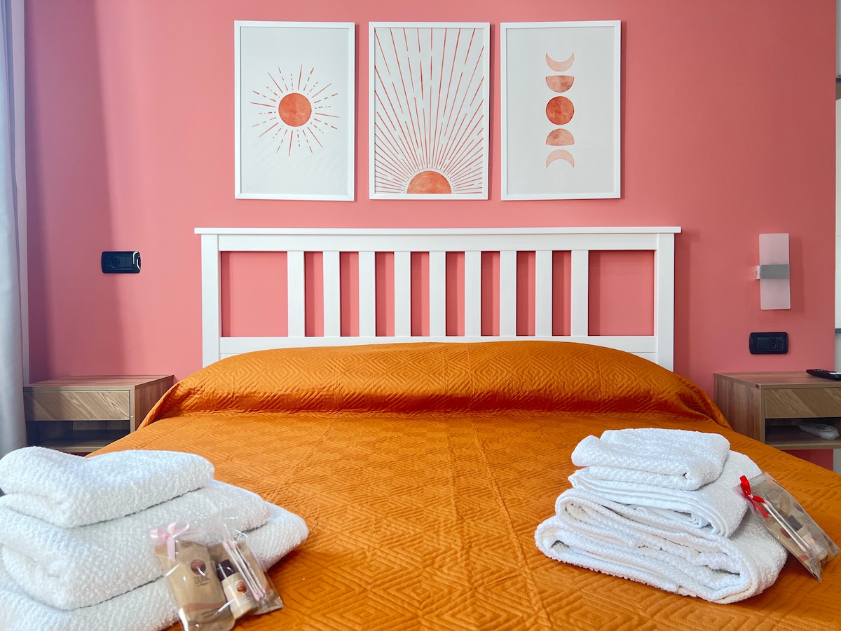 A spacious bed is adorned with an orange coverlet. Soft, white towels are neatly arranged at the foot of the bed. Three framed art pieces with sun motifs hang on the pink wall above the headboard, enhancing the room's vibrant decor.