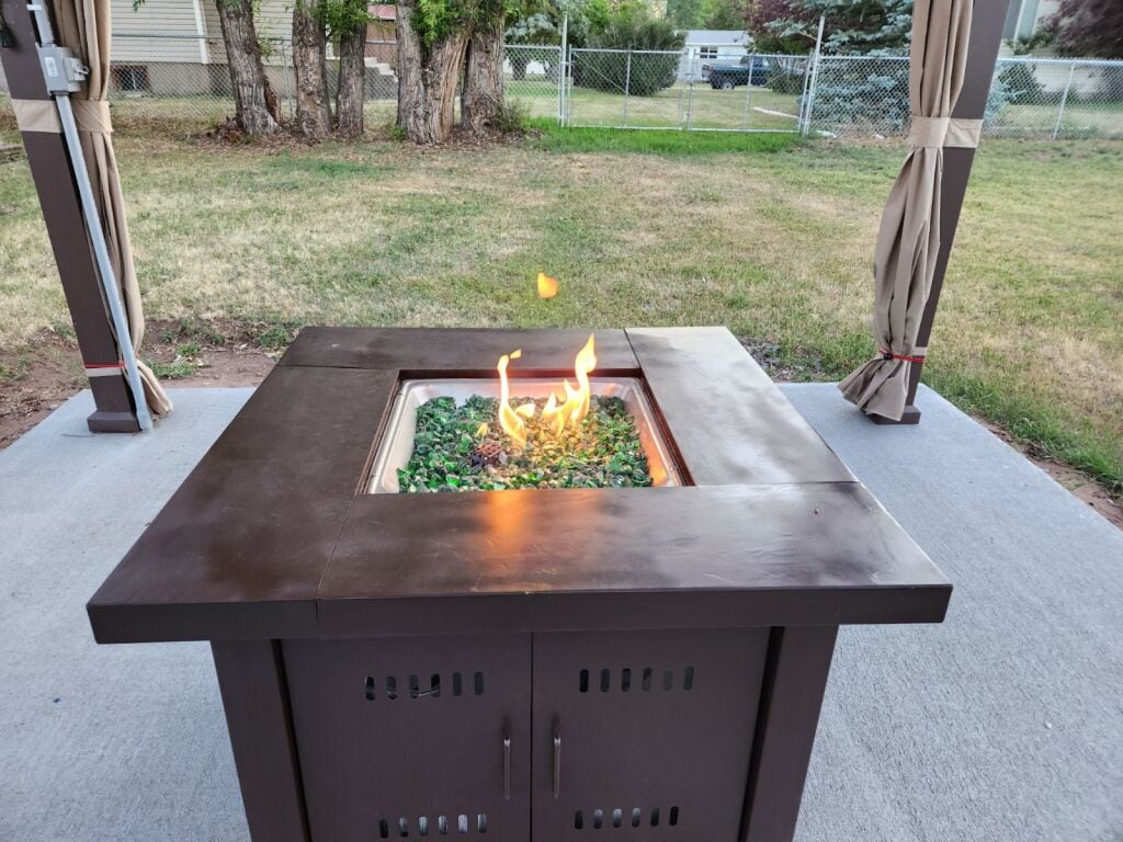 An outdoor firepit is showcased within a pergola structure, featuring a smooth tabletop and decorative stones surrounding the flames. The grassy yard is visible in the background, enhancing the relaxation space. Curtains drape alongside the pergola, contributing to a cozy atmosphere.