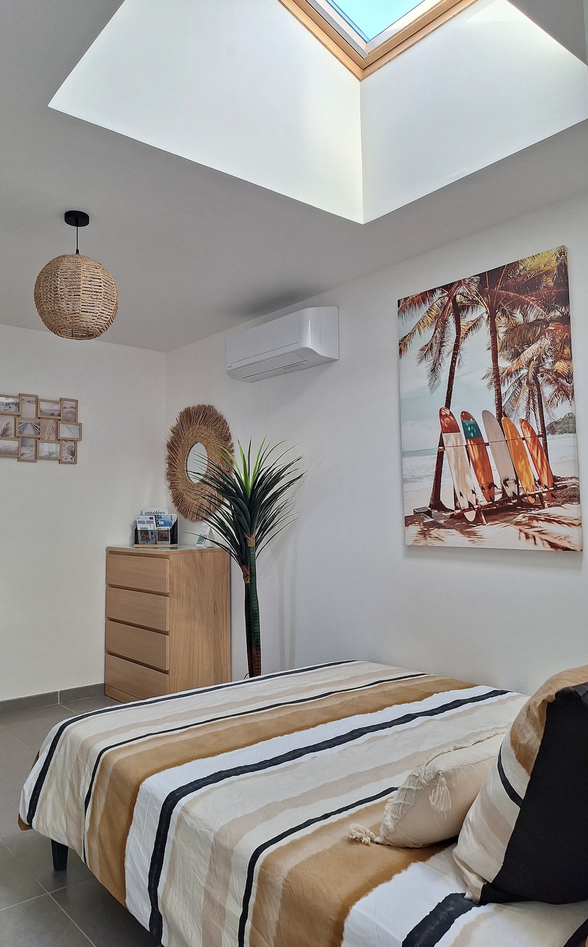 A well-lit sleeping area features a spacious bed adorned with striped bedding and a decorative cushion. A dresser is positioned against one wall, complemented by a palm tree artwork. A ceiling fan and air conditioning unit provide climate control, and natural light filters through a skylight.