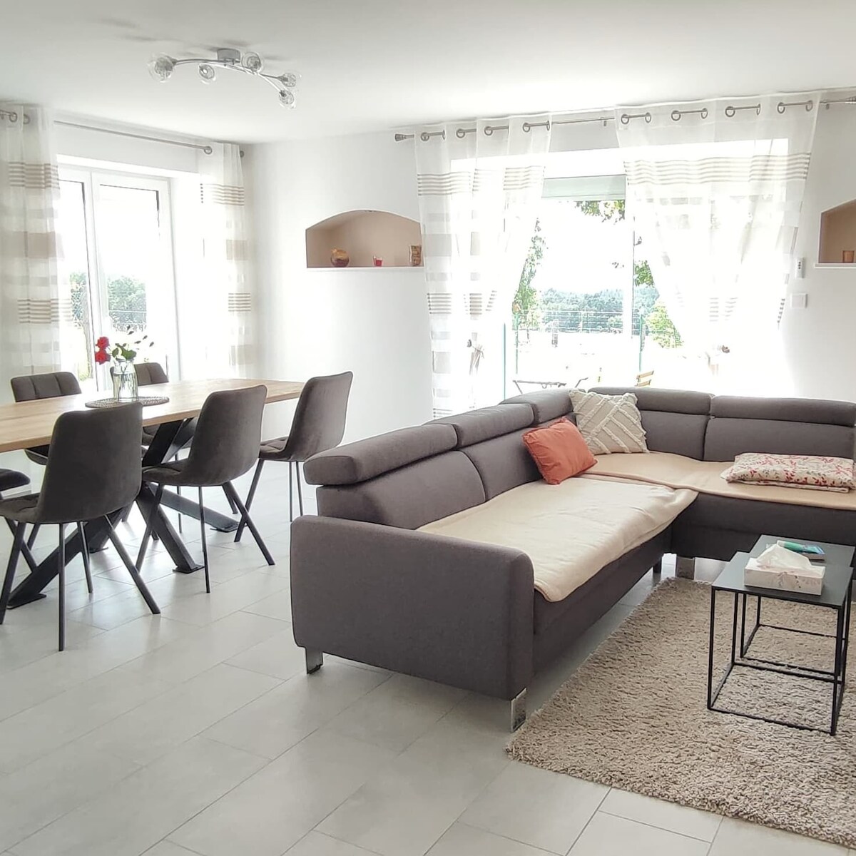 A spacious living and dining area features a comfortable sectional sofa with decorative cushions, a coffee table, and a large dining table with six chairs. Natural light enters through large windows, showcasing views of the outdoor landscape beyond. Light curtains enhance the airy atmosphere.