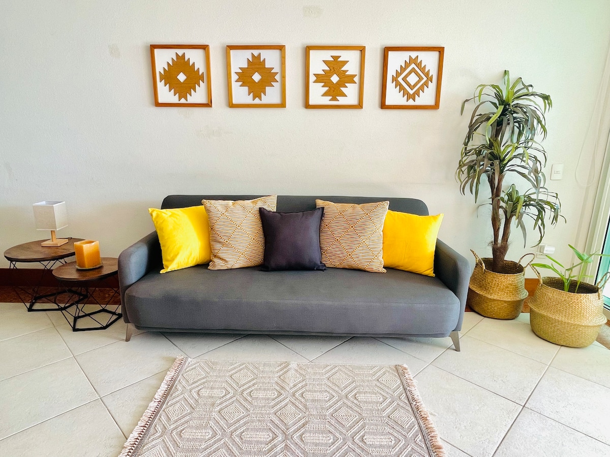 A modern living area features a gray sofa adorned with colorful cushions in yellow and black. Two patterned artworks are displayed above the sofa. A small table with a candle and a lamp is positioned nearby, accompanied by potted plants and woven baskets on the floor.