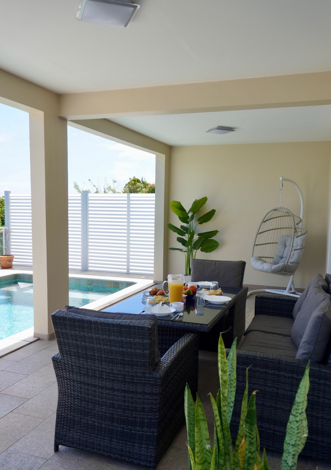 An outdoor terrace features a comfortable seating arrangement with dark wicker chairs surrounding a table set with refreshments. A tropical plant adds greenery, while a small pool is visible nearby. A hanging chair is positioned beside a privacy screen, enhancing the serene environment.