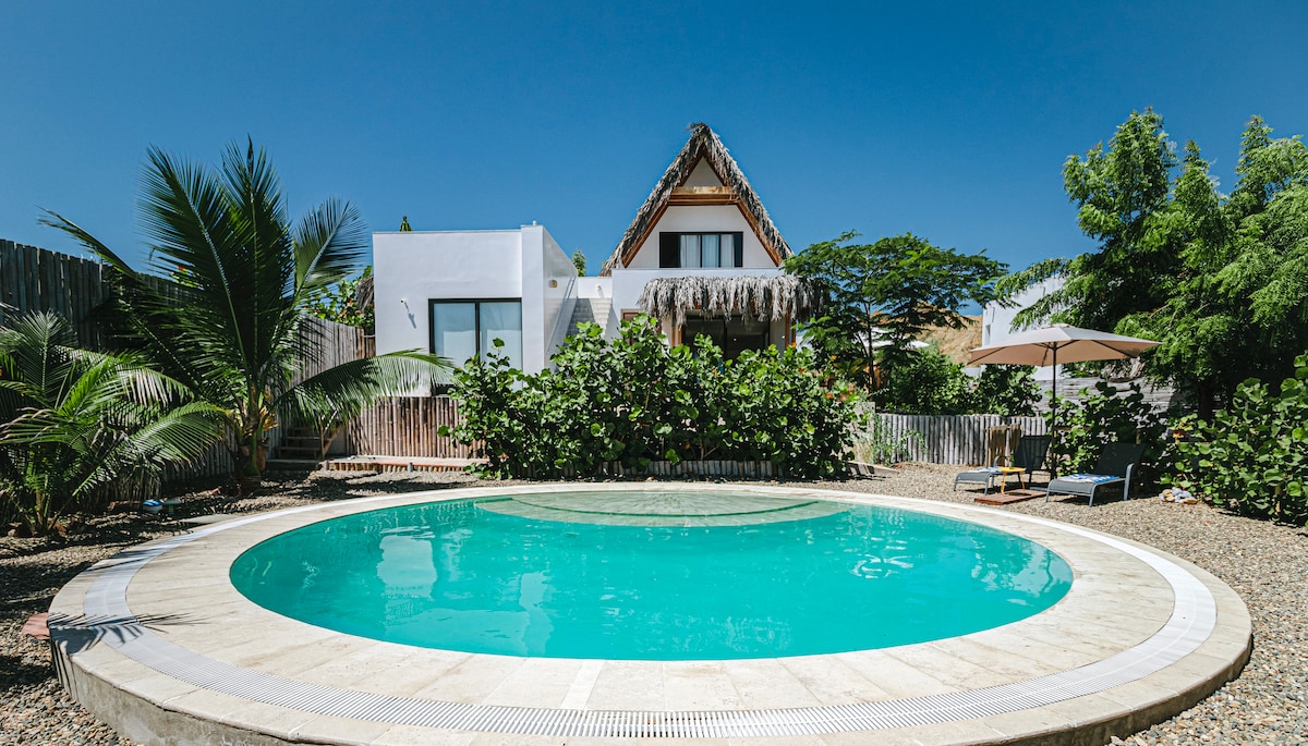 A circular pool sits surrounded by lush greenery and tropical plants. The house with a thatched roof is visible in the background, featuring large windows and a welcoming terrace. Sun loungers are positioned to the side, offering a relaxing outdoor space under the clear blue sky.