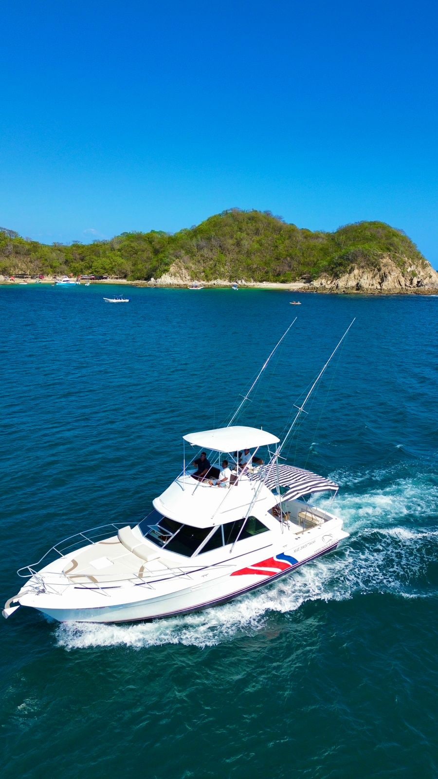 A white motorboat is seen navigating through clear blue waters, with sunlight reflecting off the surface. The vessel features a spacious upper deck and fishing rods mounted at the rear. A lush, green coastline is visible in the background.