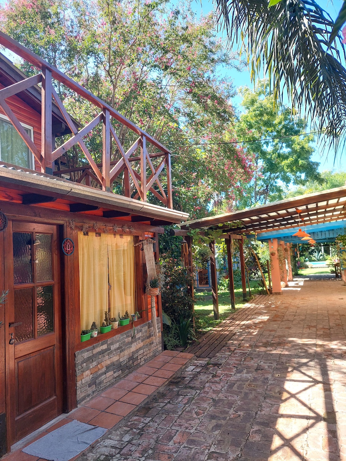 A charming pathway is lined with lush greenery and flowering bushes, leading to a wooden entrance adorned with elegant curtains. The area is sunlit, showcasing a covered space with seating and colorful umbrellas, inviting relaxation in a tranquil outdoor setting.