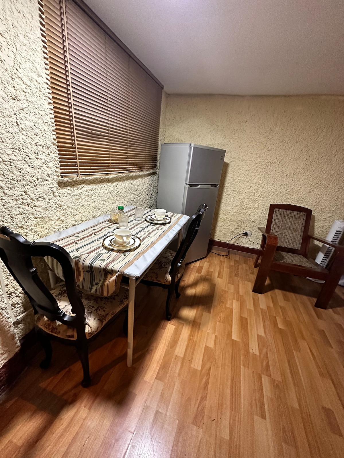 A small dining area features a table set for two, complemented by elegant chairs. A compact refrigerator is positioned nearby against a textured wall, while natural light filters in through a window covered with wooden blinds. The flooring is a warm wood tone.