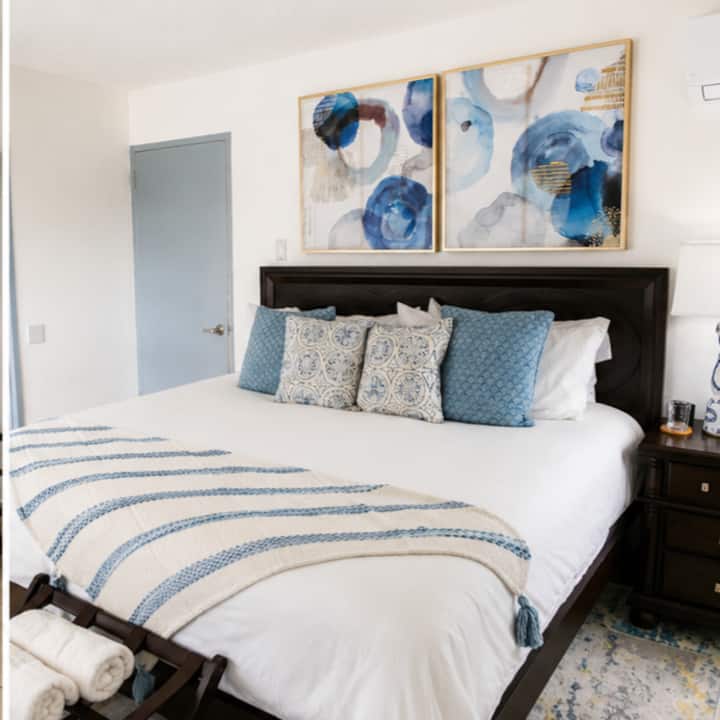 Featuring a plush king-size bed dressed in crisp white linens with coastal-inspired décor, the space offers a peaceful atmosphere for restful nights. Soft blue tones, elegant artwork, and warm wood furnishings create a refined coastal feel.