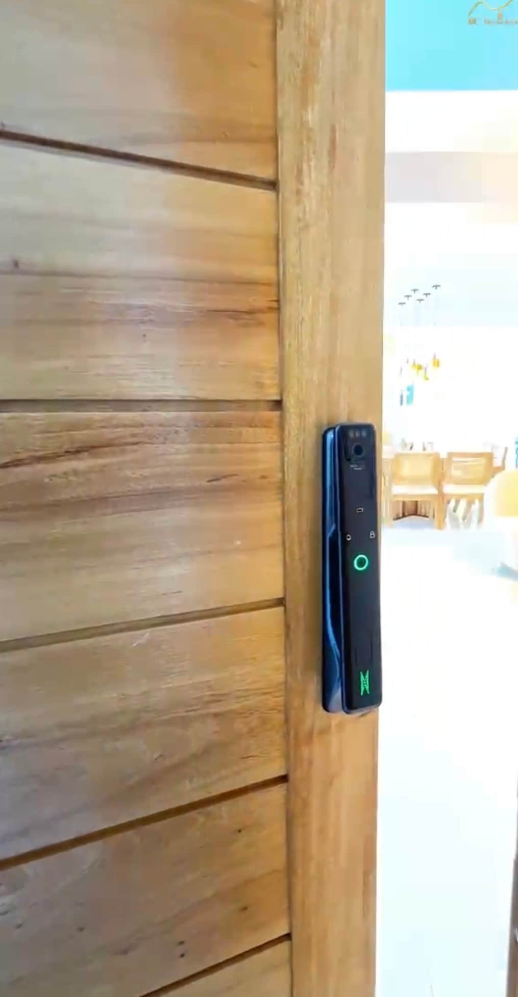 A modern keyless entry system is installed on a wooden door, showcasing the natural texture of the wood. The door is partially open, revealing a well-lit living area in the background with wooden furnishings and a welcoming ambiance.