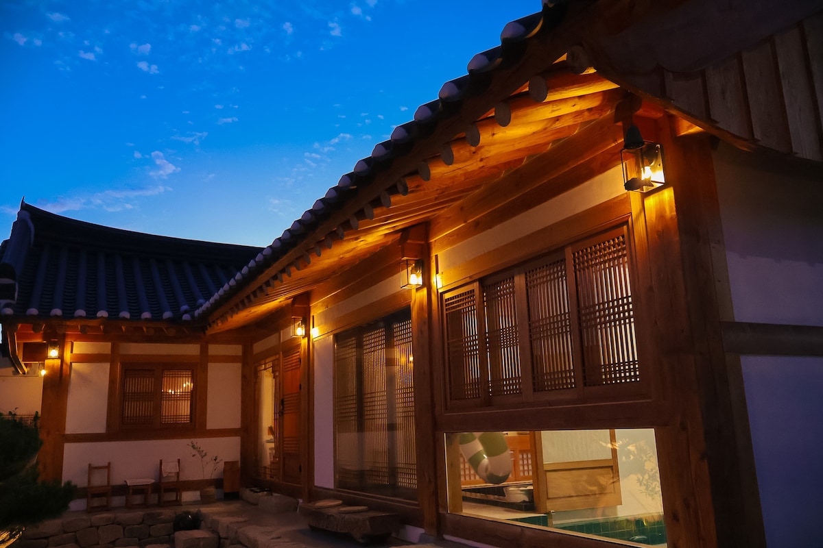 The exterior of a traditional Hanok features warm lighting that highlights the wooden structure's intricate design. A serene courtyard is visible, framed by lush greenery and stone pathways, with a glimpse of the indoor swimming pool seen through large glass windows.