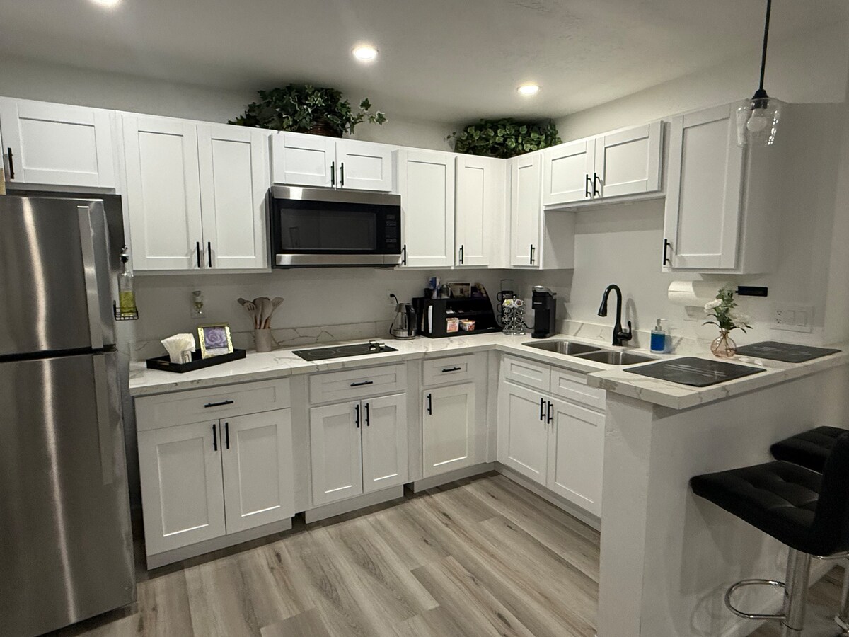A modern kitchen features white cabinetry and stainless steel appliances. A microwave and coffee station are visible, alongside a sink and a stylish faucet. The countertops are adorned with minimal decor, while plants add a touch of greenery to the space. Bar stools are positioned near the countertop.