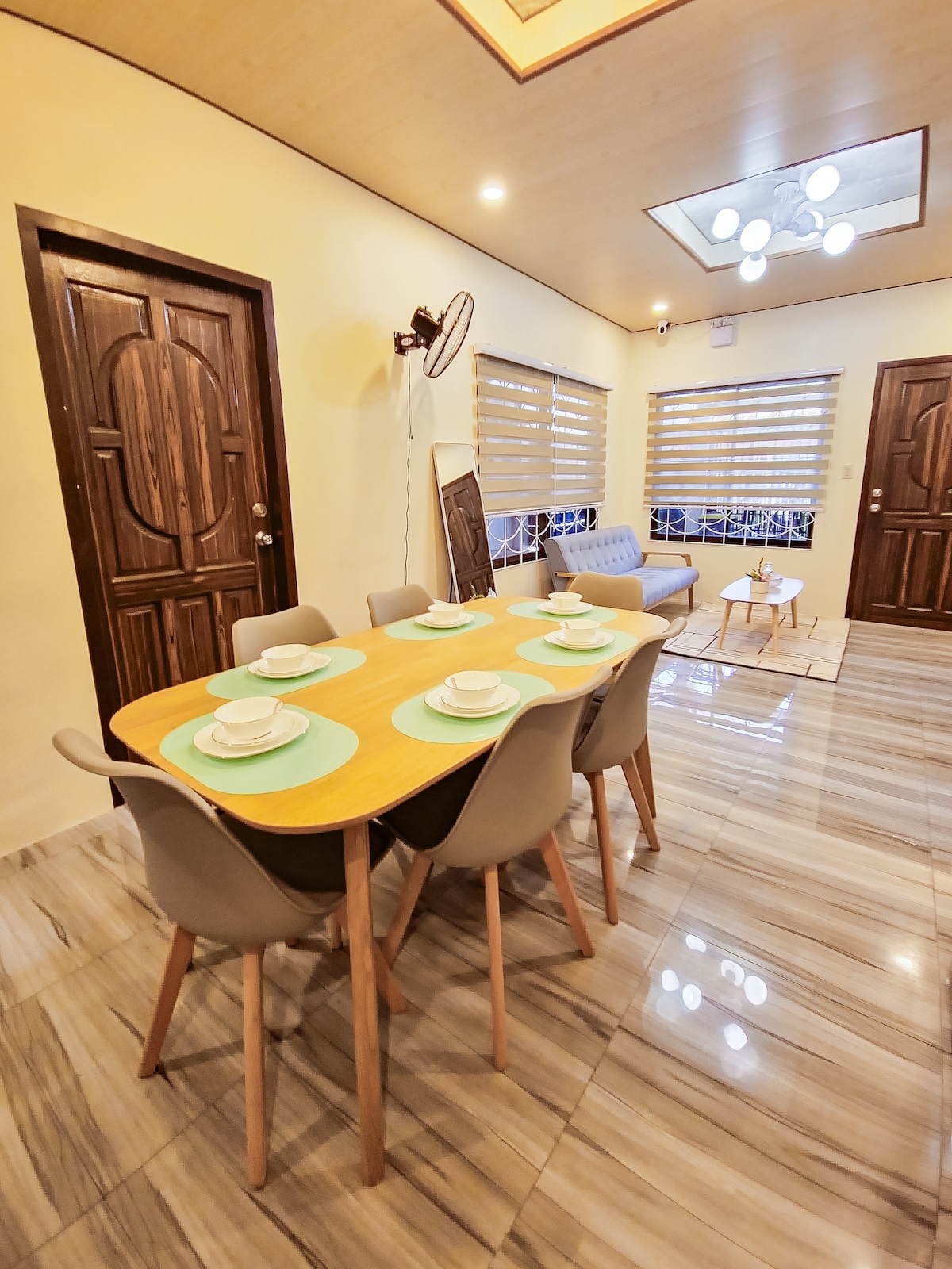 A dining area features a wooden table set for six, complemented by light grey chairs. Natural light enters through large windows adorned with blinds. A cozy seating arrangement is visible in the background, adding to the room's inviting atmosphere.