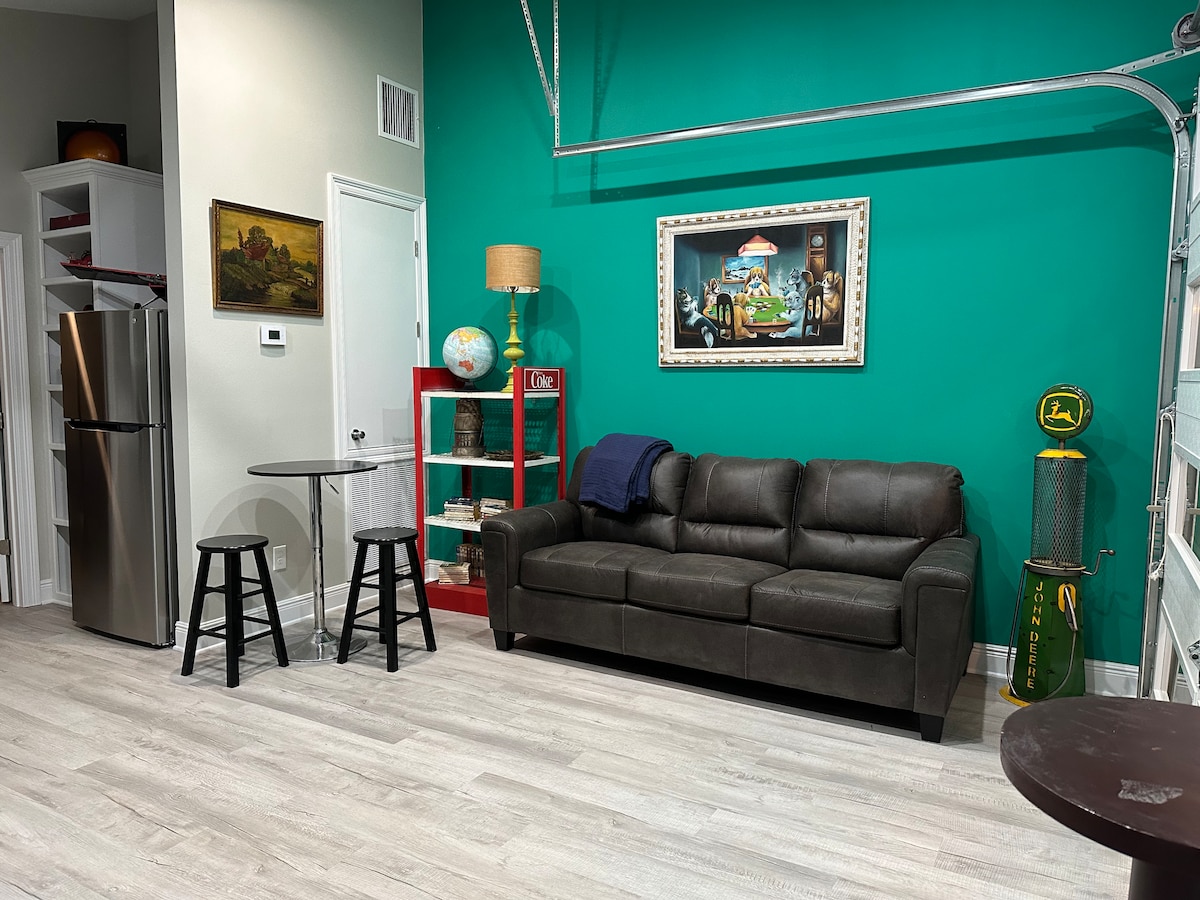 A vibrant green wall complements a cozy living space featuring a dark fabric sofa. A small round dining table with two black stools is positioned nearby. A vintage globe and artwork add character to the area, while a stainless steel refrigerator is visible in the background.