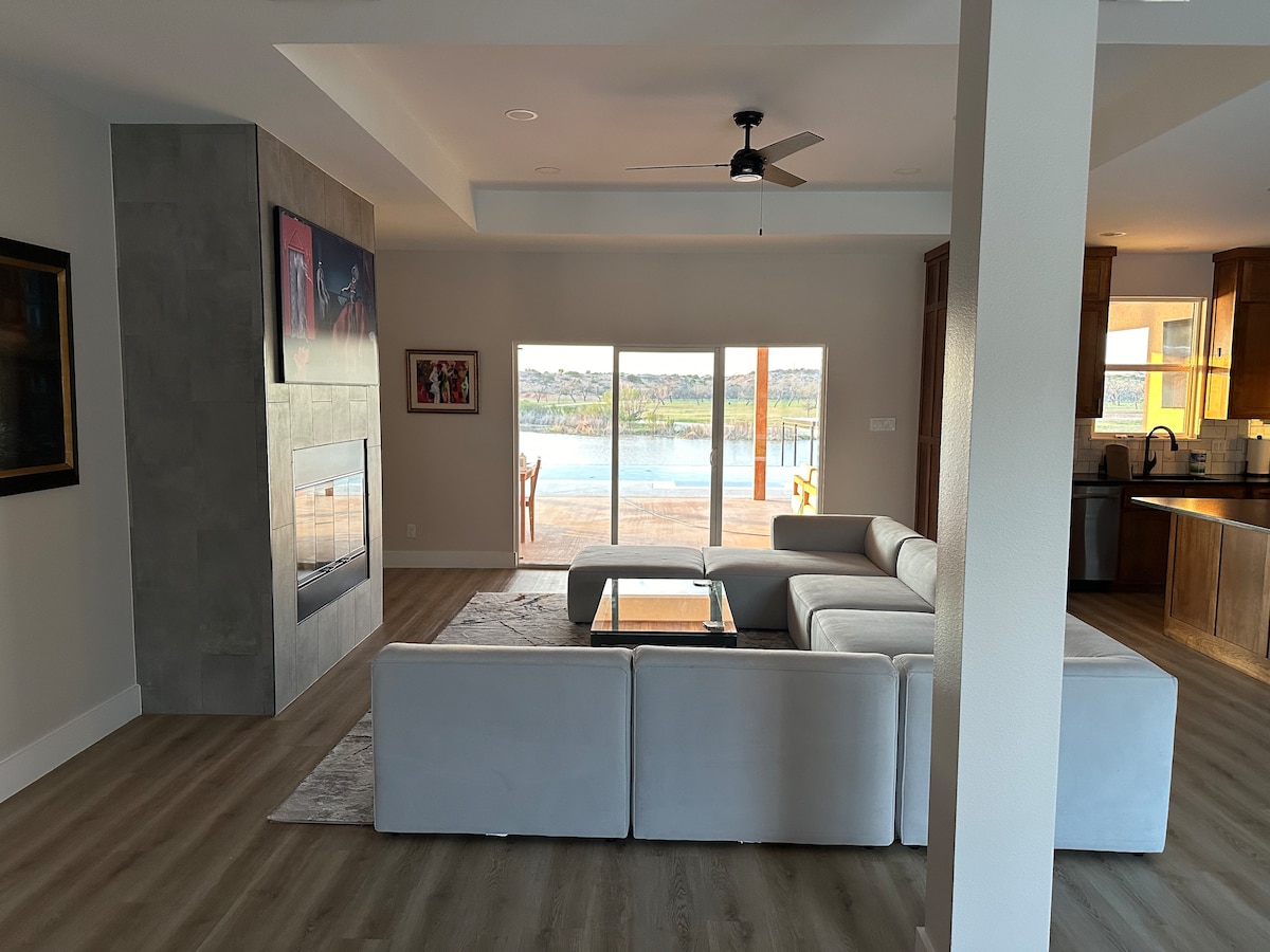 A spacious living area features a modern sectional sofa facing a sleek, wall-mounted fireplace. Large glass doors provide an unobstructed view of the lake and outdoor deck. Natural light fills the room, highlighting the neutral decor and light wood flooring.
