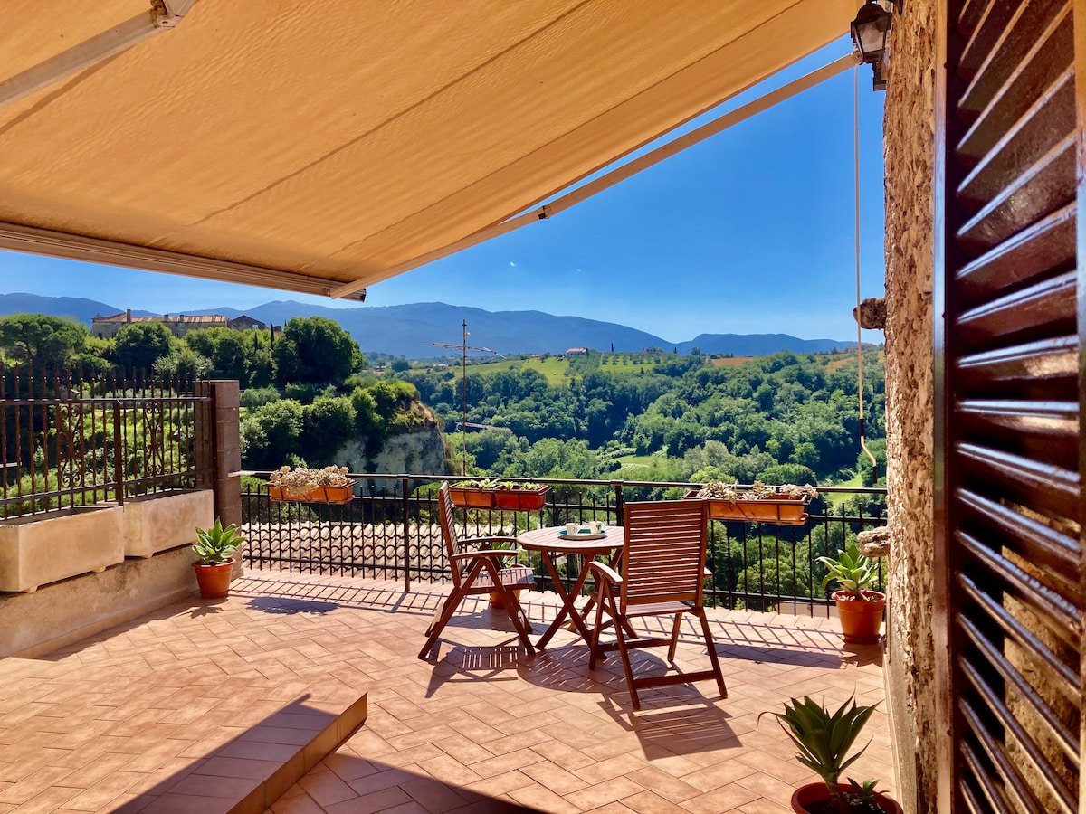 A private terrace is showcased, featuring a small round table and two wooden chairs, overlooking a lush valley. The space is enhanced by potted plants and a retractable awning, providing shade and comfort while enjoying captivating views of the surrounding landscape.