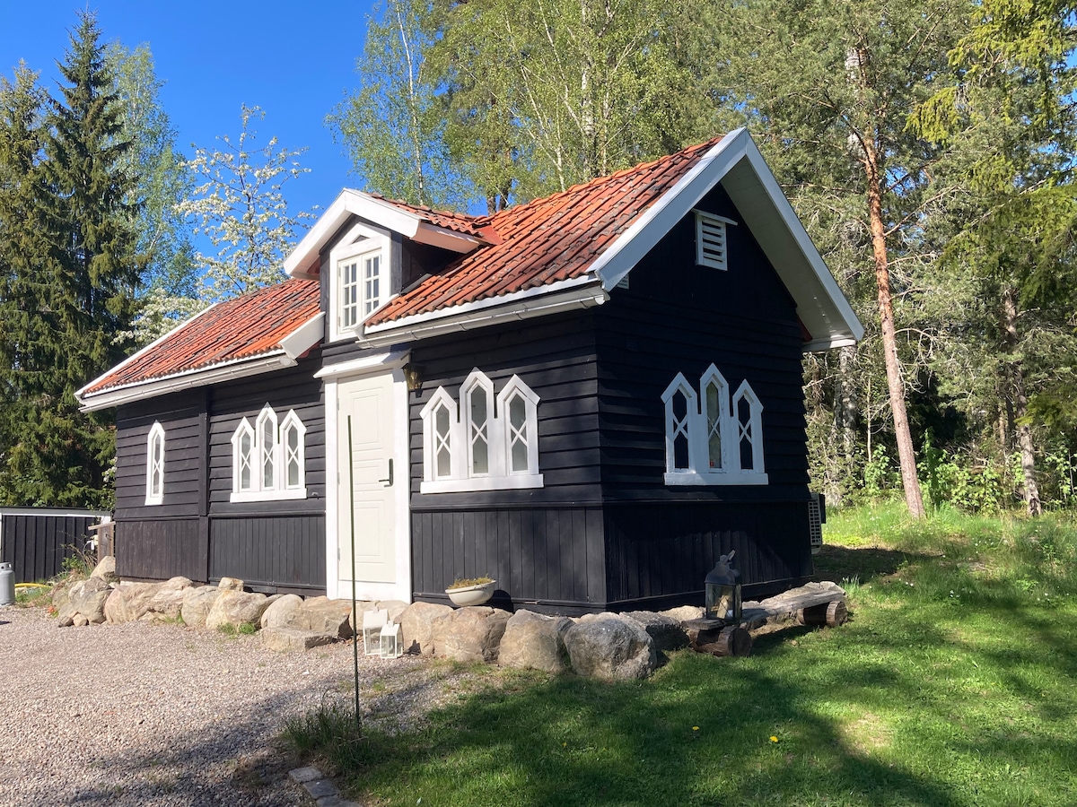 The charming exterior of the cabin features a dark wood facade complemented by white, arched windows. A welcoming front door is positioned centrally, framed by a neatly maintained gravel area. Lush greenery surrounds the cabin, enhancing its tranquil setting.
