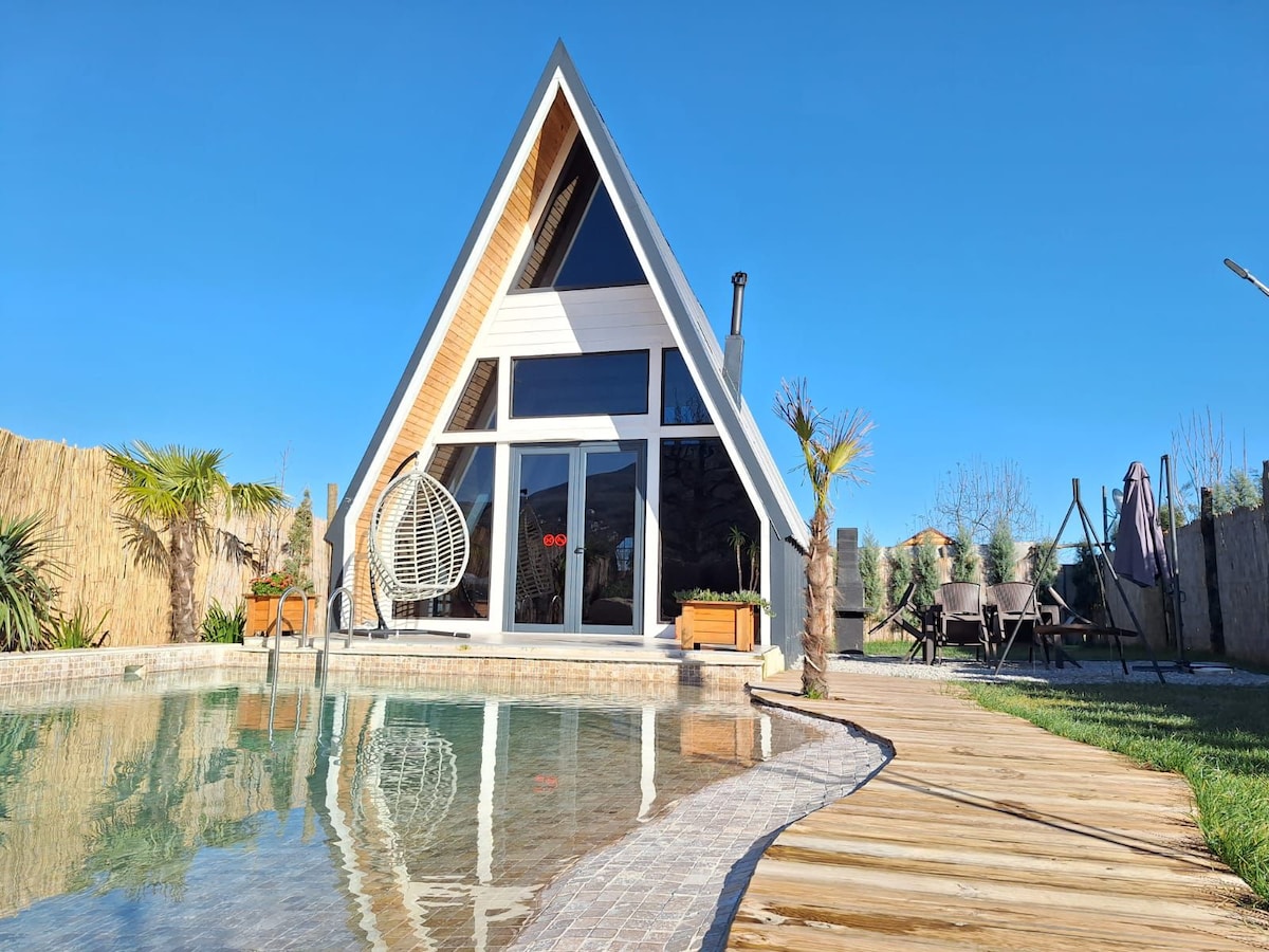 A modern A-frame house features large glass windows and a circular hanging chair. The adjacent pool reflects the clear blue sky. Surrounding greenery and wooden pathways lead to a seating area with lounge chairs, contributing to the serene outdoor setting.