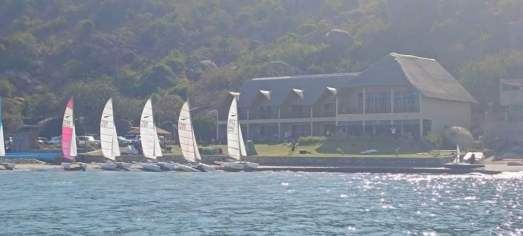 A charming beachfront building is visible, featuring a thatched roof and multiple windows across the upper level. Various sailboats are lined up along the shore, their white sails contrasting with the lush green landscape in the background.