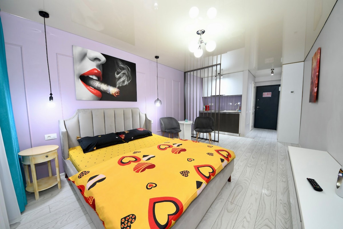 A modern sleeping area features a spacious bed with a brightly colored blanket adorned with heart motifs. A bold wall art piece of a woman's lips is displayed above the bed. Soft lighting is provided by pendant lamps, and glimpses of the kitchen area are visible in the background.