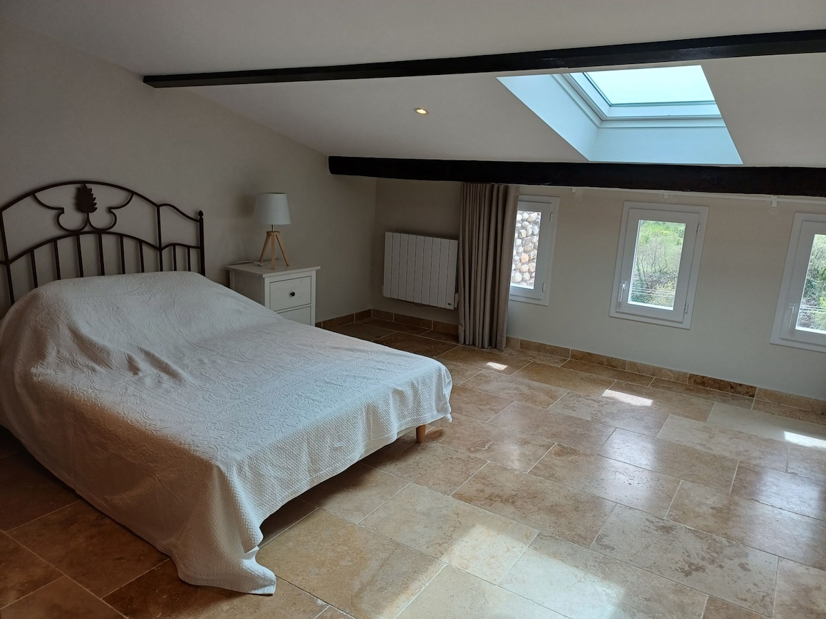 A spacious bedroom is illuminated by natural light streaming through a skylight and two small windows. A simple metal bed frame supports a neatly made bed. A bedside table and a radiator are positioned adjacent to the bed, with stone flooring adding a warm touch.