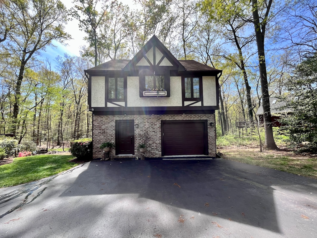 The exterior of a two-story Tudor-style home is framed by tall trees, showcasing a brick façade and a decorative upper section. A single garage door complements the structure, while a paved driveway leads up to the entrance. Natural light filters through the surrounding foliage.