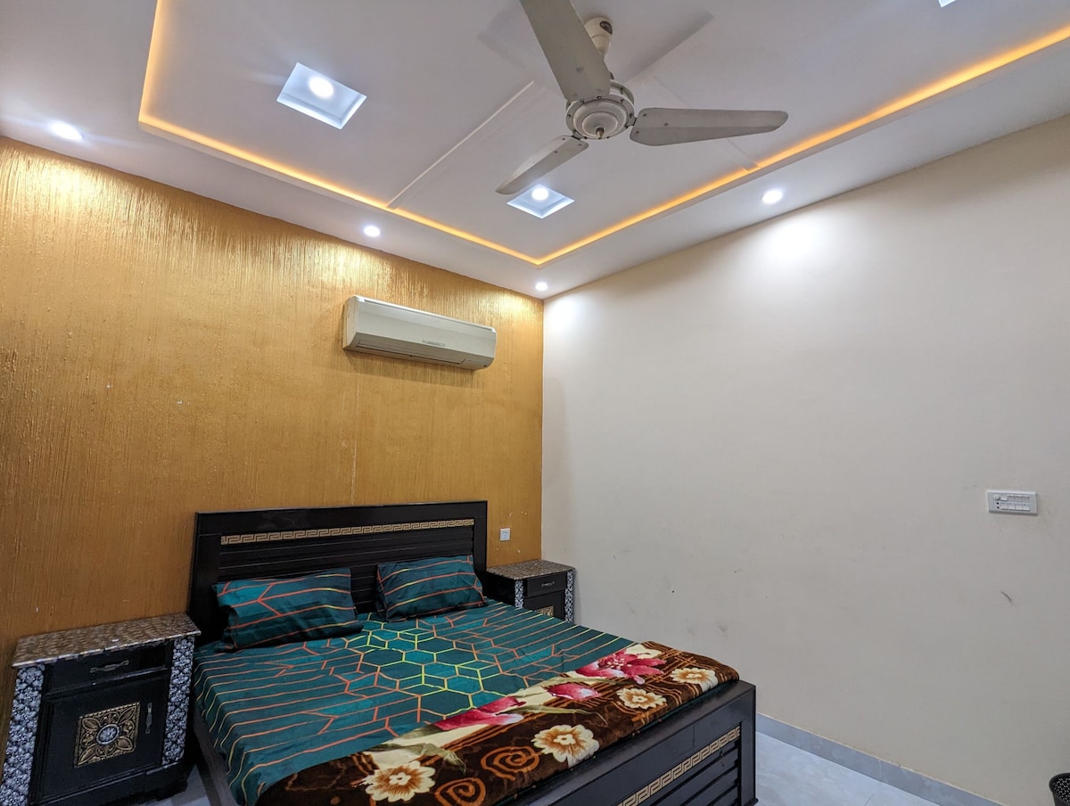 A spacious bedroom features a stylish black bed with a colorful patterned comforter. The walls are adorned with a textured golden finish, and ambient lighting is provided by recessed ceiling lights. A ceiling fan offers ventilation, while an air conditioning unit is mounted above the bed.