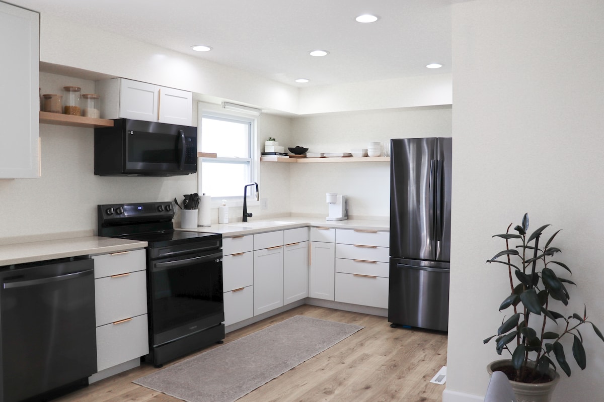 A modern kitchen features sleek cabinetry with neutral tones and wooden accents. New appliances, including a black stove and refrigerator, are complemented by natural light from a nearby window. A subtle rug adds warmth, while potted greenery enhances the calming atmosphere.