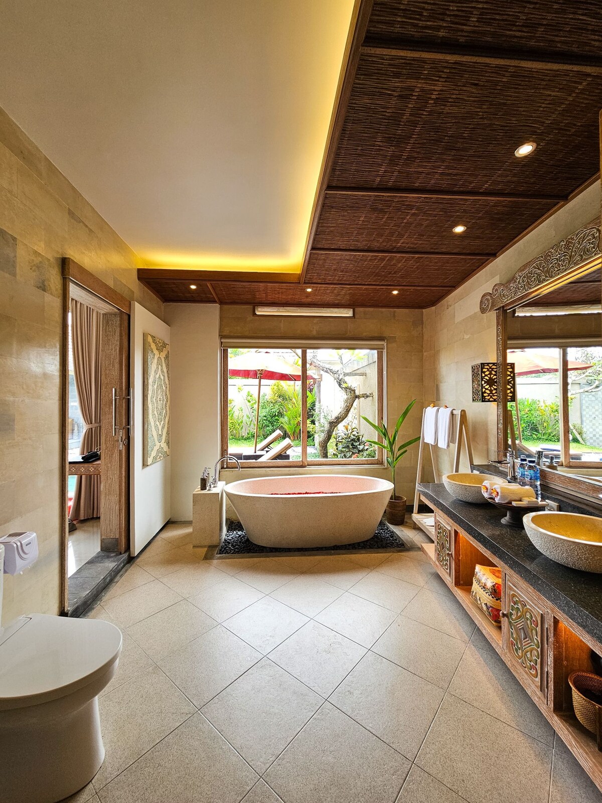 A luxurious bathroom features a freestanding bathtub positioned near a large window overlooking greenery. Natural light fills the space, highlighting the elegant stone finishes. Two vessel sinks are placed on a dark countertop, while bamboo accents add a touch of warmth to the room.
