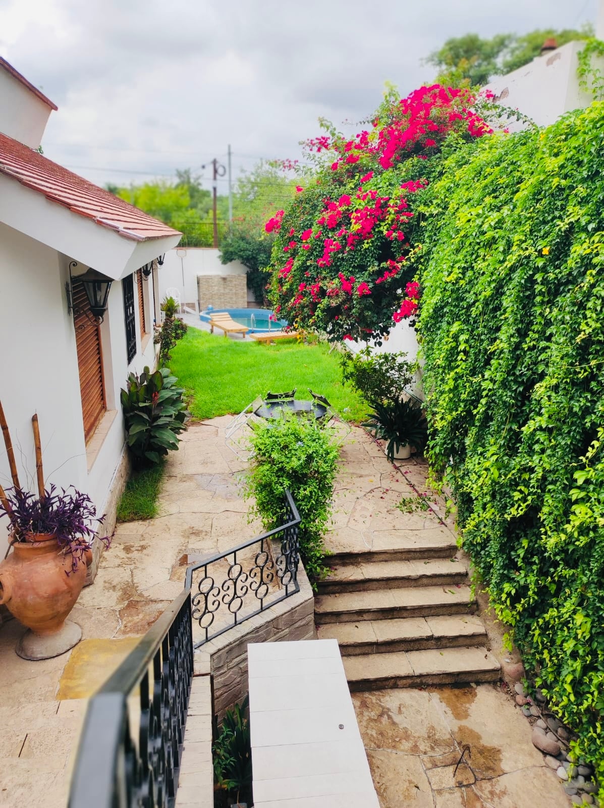 A view of the garden showcases lush greenery and vibrant flowers, with a small pool nestled in the background. Stone steps lead from the house to the lawn, framed by climbing plants and colorful blooms that enhance the outdoor space.