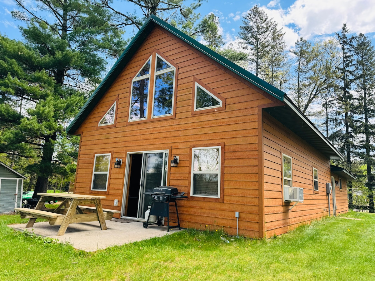 The cabin exterior features a wooden facade with large triangular windows, allowing natural light to brighten the interior. A picnic table and a grill are positioned on the patio, surrounded by lush grass and tall trees in the background.
