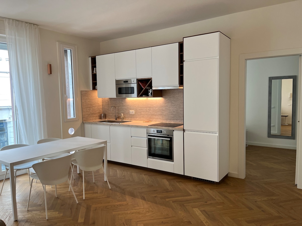 An open kitchen space is presented, featuring white cabinetry and modern appliances. Under-cabinet lighting subtly illuminates the countertop. A dining table with white chairs is positioned nearby, while the warm wood floor adds texture. A distant section offers a glimpse of additional living space.
