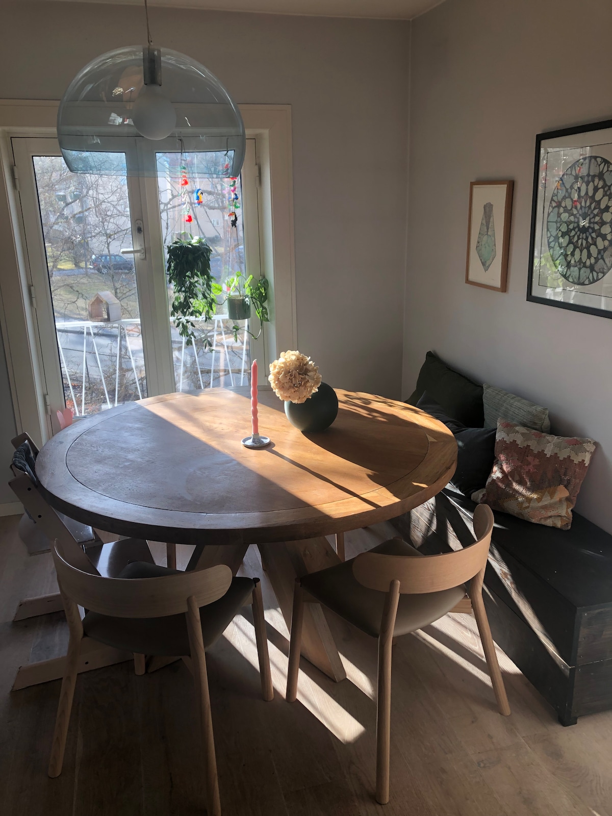 A round wooden dining table is illuminated by sunlight, surrounded by four wooden chairs. A simple vase with flowers is positioned at the center, accompanied by a single candle. A cozy seating area with cushions is visible in the background, framed by large glass doors offering views of the outdoors.