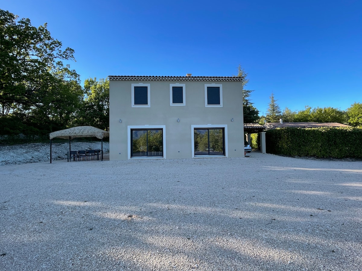 The exterior of the house is shown, featuring a two-story structure with multiple large windows and a covered seating area. A spacious gravel area surrounds the property, accompanied by greenery in the background, contributing to a serene environment.