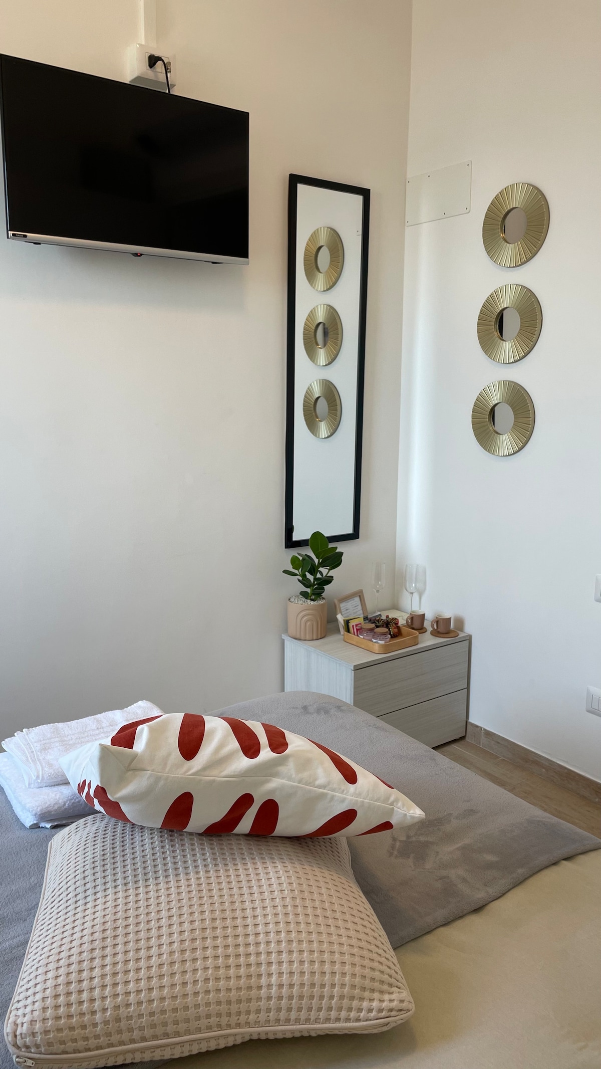 A well-lit bedroom is presented, featuring a flat-screen television mounted on the wall. A mirror with a sleek frame is accompanied by three decorative wall mirrors. A nightstand displays a small plant and some decor, alongside neatly arranged pillows on the bed.