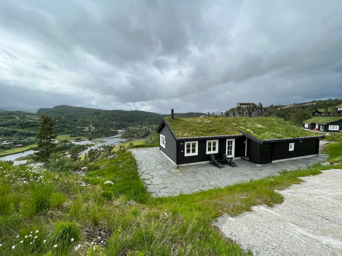 A modern black cabin is set against a backdrop of rolling hills and a winding river. The stone patio features two lounge chairs, inviting relaxation. The cabin's green roof seamlessly integrates with the natural surroundings, enhancing its tranquil position in the landscape.