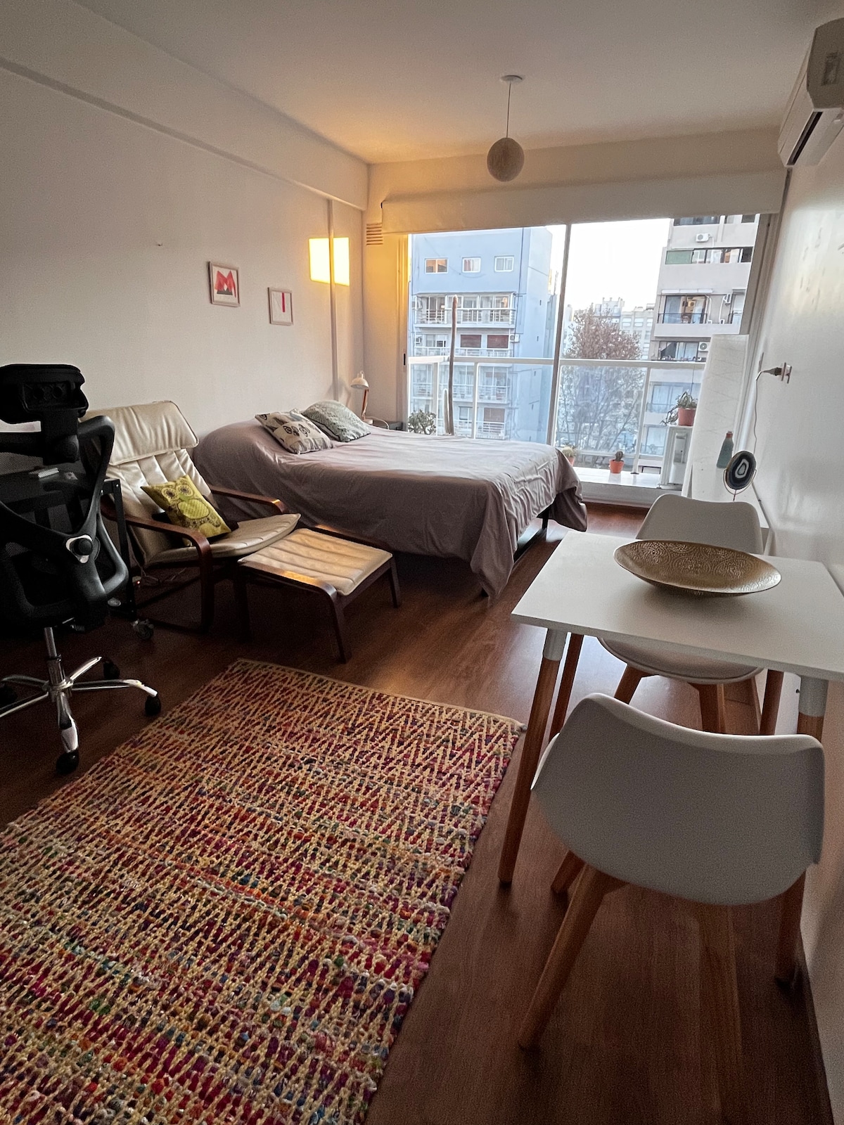 A studio apartment is displayed, featuring a spacious bed with a grey coverlet and a small dining table with two chairs. A cozy armchair and desk are positioned near the window, which offers views of the surrounding buildings. A colorful area rug adds texture to the wooden floor.