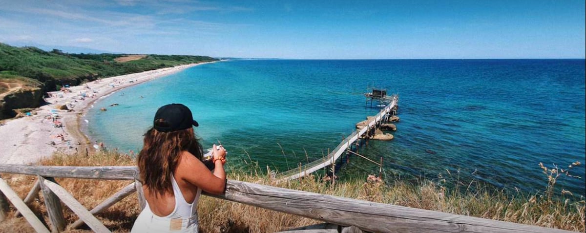 A scenic view of a beach with soft sand and clear blue waters is presented. A wooden pier extends into the sea, while sunbathers occupy the shoreline. Lush greenery frames the coastline, and a person is seen leaning against a wooden railing, enjoying the landscape.