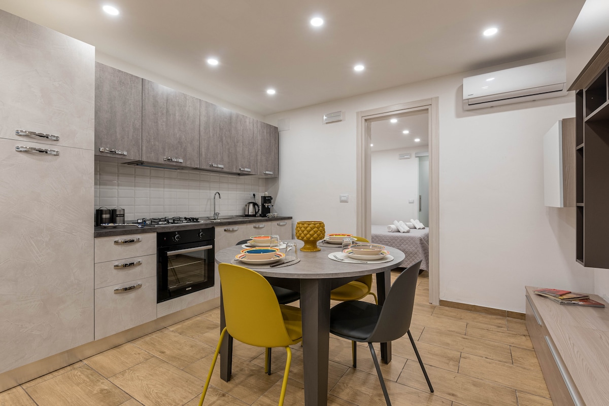 A modern kitchen area features a round table with four chairs, complemented by light-colored cabinetry. A built-in oven and stovetop are visible, along with a coffee maker. Soft lighting illuminates the space, creating a welcoming atmosphere that connects to the adjacent bedroom.