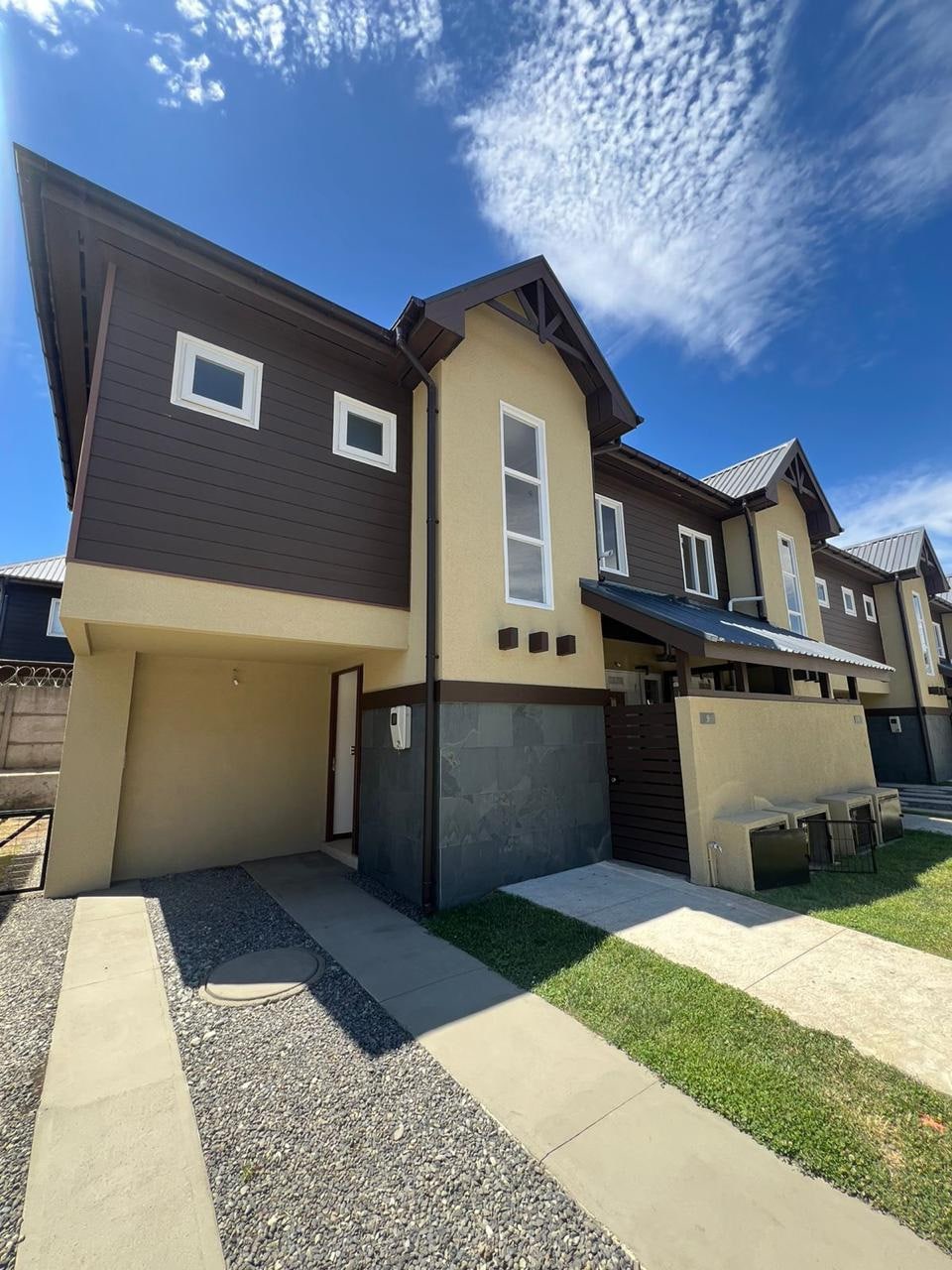The exterior of the two-story house displays a modern design with a combination of light and dark materials. A well-maintained pathway leads to the entrance, bordered by a small lawn. The clear blue sky complements the earthy tones of the building.