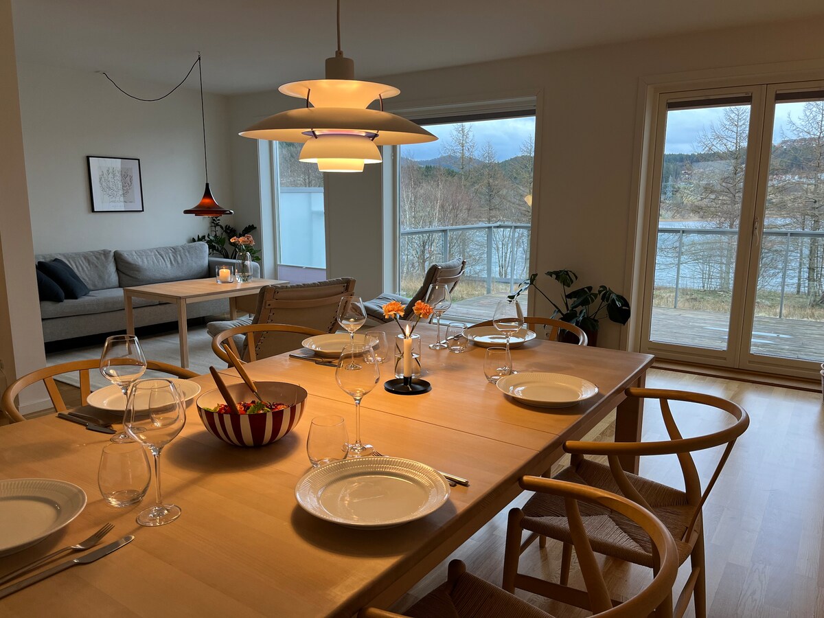 A dining table is set with plates and glasses, inviting a communal dining experience. Soft natural light enters through large windows, illuminating a cozy living area with a sofa in the background. A scenic view of the surrounding landscape is visible through the glass doors.
