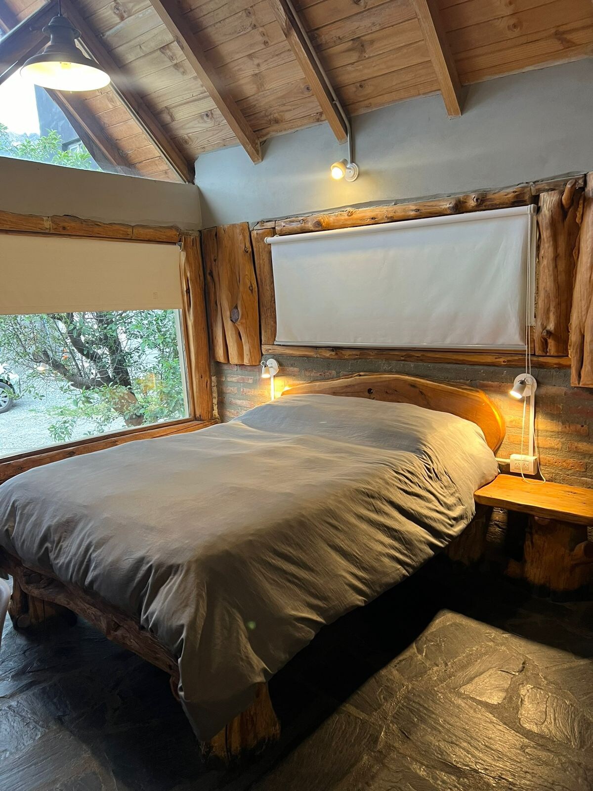 A cozy sleeping area is presented with a spacious bed, featuring a neutral-toned duvet and wooden accents. Natural light enters through large windows, showcasing the surrounding greenery. The ceiling is adorned with exposed beams, adding to the rustic charm of the space.