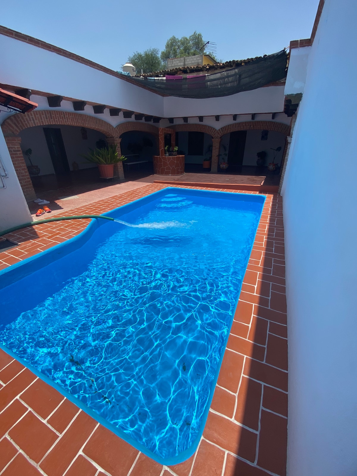 A refreshing swimming pool is situated in a tiled courtyard, surrounded by arched columns. Sunlight reflects off the clear blue water, creating a serene atmosphere. The pool is visible from multiple angles, enhancing the openness of the space, which includes shaded areas along the perimeter.