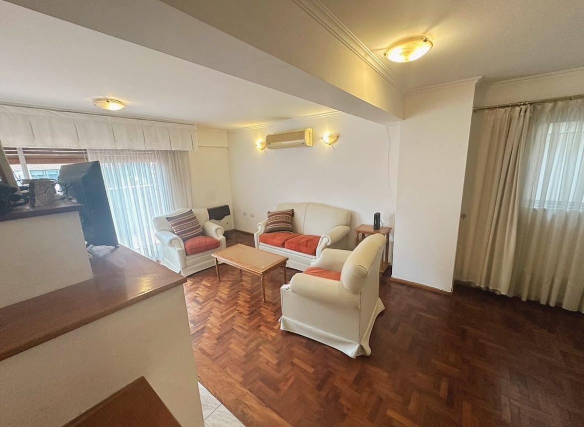A bright living area features a pair of comfortable sofas with accent pillows, positioned around a wooden coffee table. Natural light filters through large windows covered by sheer curtains, illuminating the polished wooden floor and creating a welcoming space.