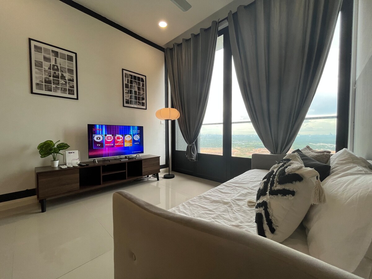The living area features a spacious day bed adorned with clean linens, complemented by a stylish black and white cushion. A 43" LED Smart TV is positioned on a wooden media console, with natural light filtering through large windows draped in charcoal curtains.