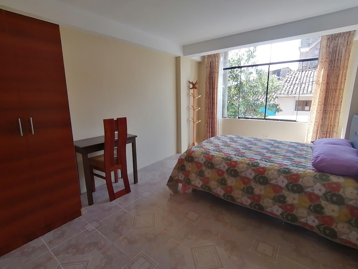 A spacious bedroom features light-colored walls and large windows that allow natural light to enter. A colorful patterned bedspread covers the bed, while a simple wooden desk and chair are positioned nearby. A wardrobe stands against the wall, providing additional storage.