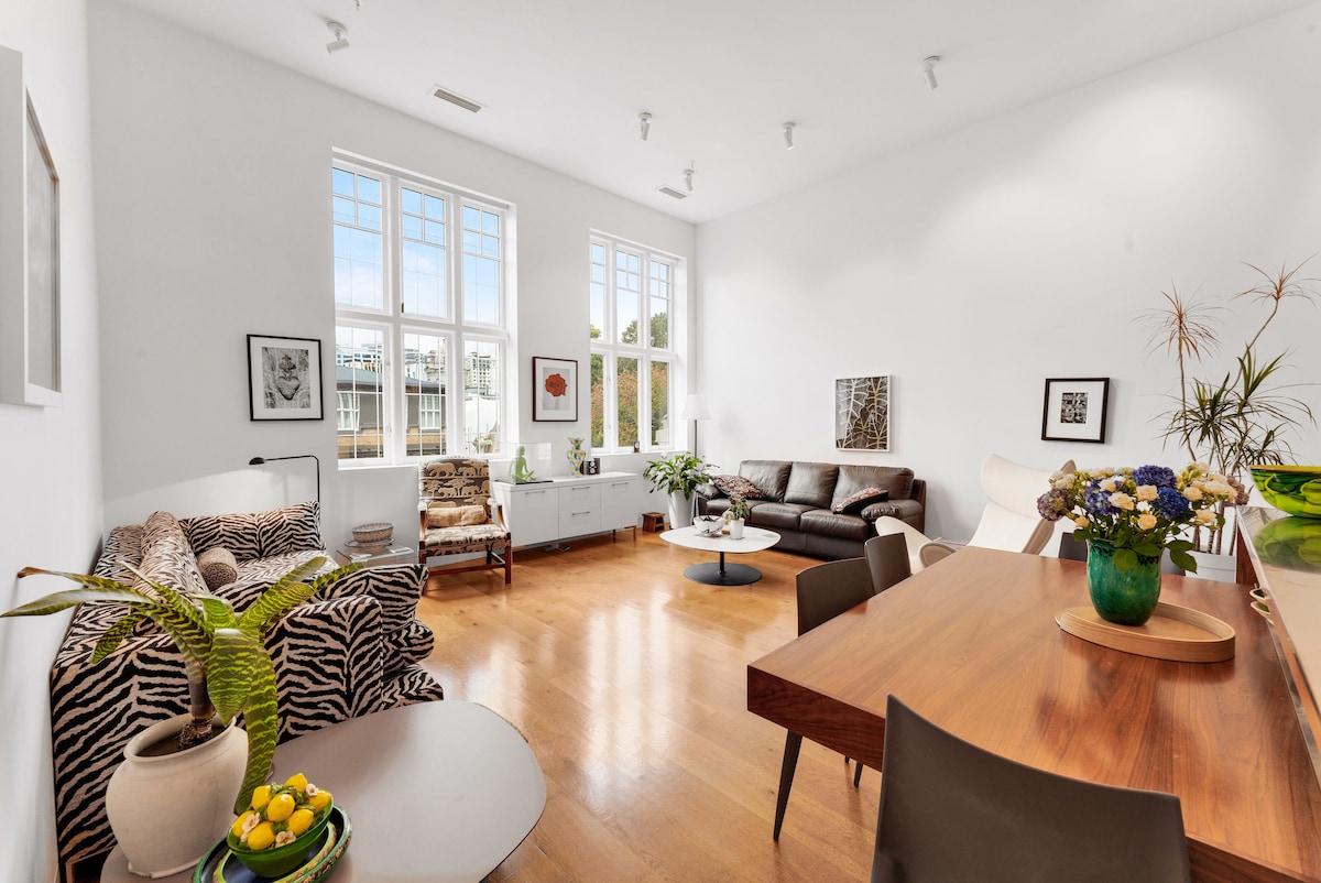 A spacious living area includes large windows allowing abundant natural light. The room features a mix of stylish seating, a dining table, and decorative elements such as plants and flowers. Polished wood flooring contributes to the modern aesthetic and comfortable atmosphere.