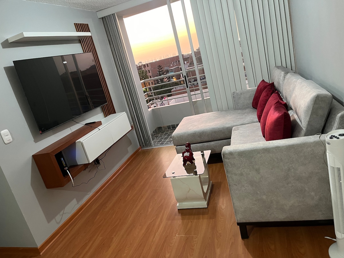 A comfortable living area is presented with a cozy gray sectional sofa adorned with red accent pillows. A wall-mounted television is visible above a sleek media console. A glass coffee table rests on the wooden floor, complementing the room's window that offers a view of the sunset.